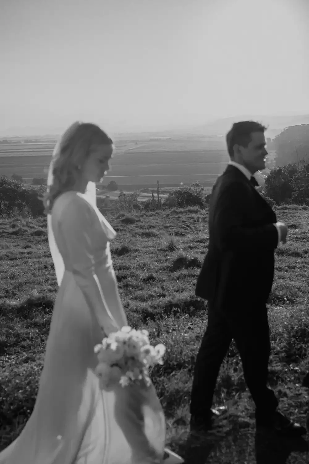 A black-and-white photo of a bride and groom walking in a countryside landscape during sunset. The bride is holding a bouquet of flowers and wearing a long dress, while the groom is dressed in a suit with a bow tie.
