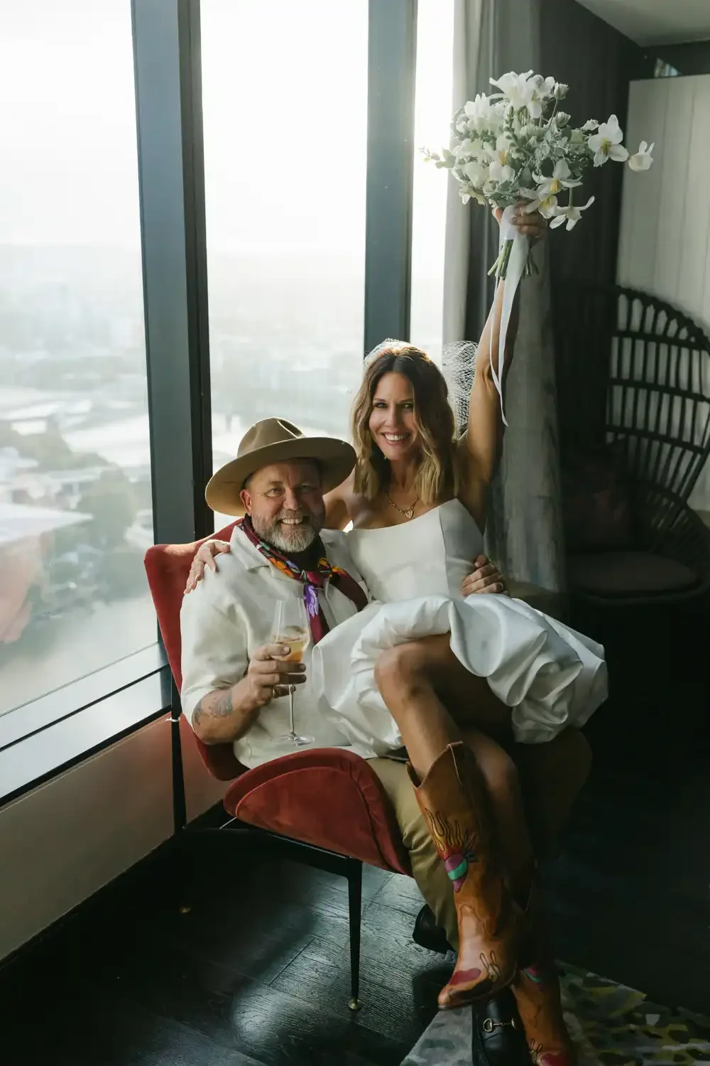 A woman in a wedding dress holding a bouquet of white flowers, sitting on the lap of a man wearing a cowboy hat, glasses, and boots, with a drink in hand, in a modern room with large windows and city view.