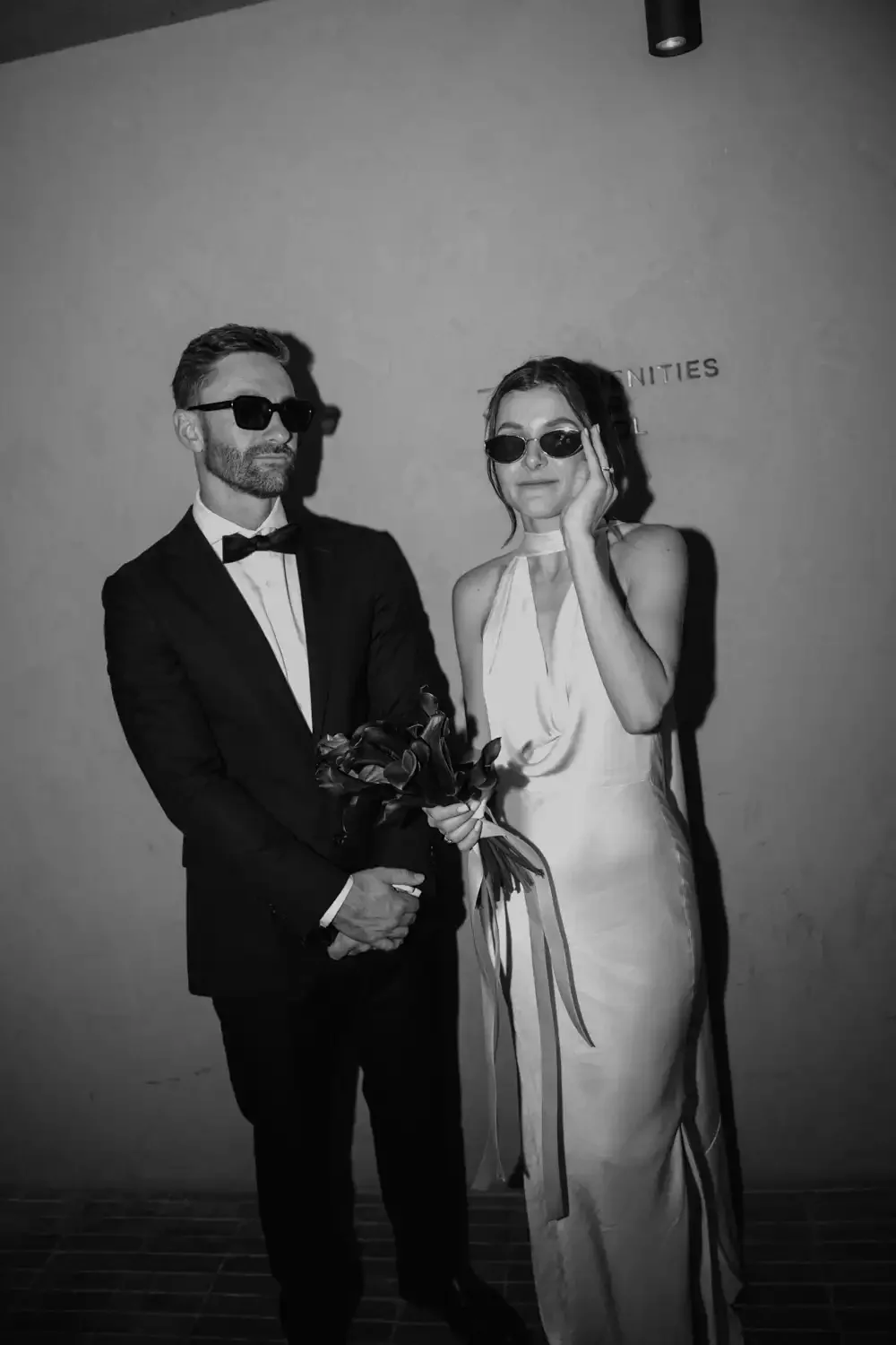 A man in a tuxedo and a woman in a white dress posing together, both wearing sunglasses. The man is holding a bouquet of flowers. The background appears to be indoors with partial text visible on the wall.