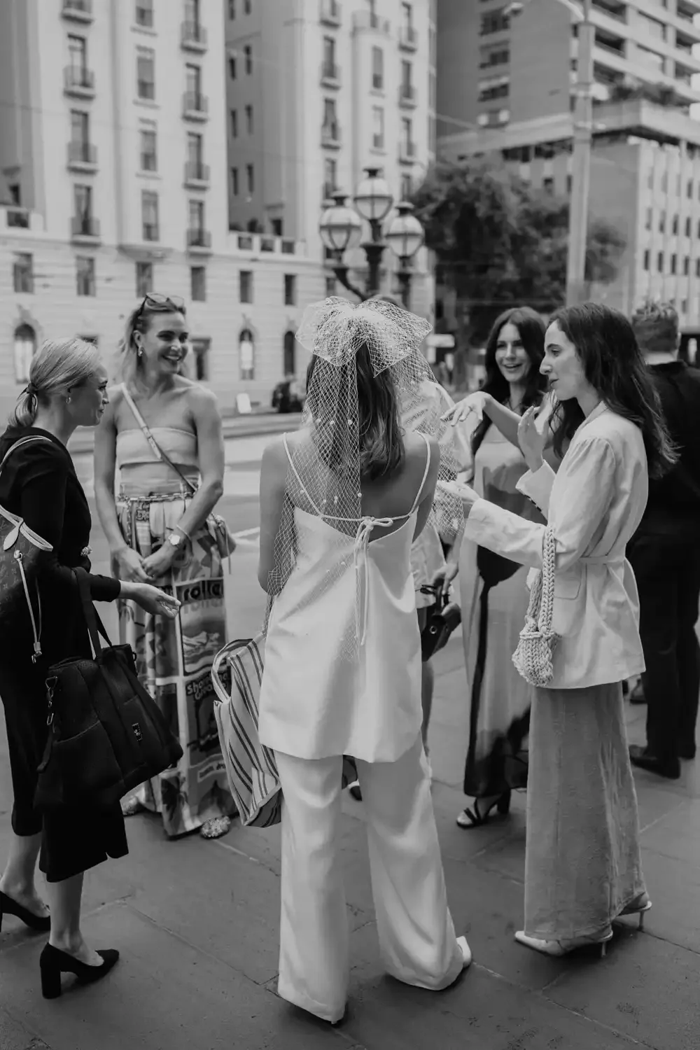 Group of women chatting on a city sidewalk, one woman wearing a veil and dress, suggestive of a bridal event or celebration.