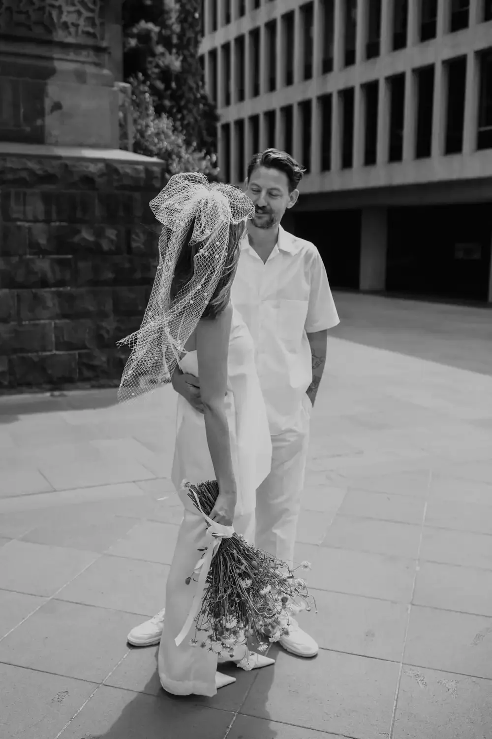 A man and a woman standing outdoors in an urban area, sharing a moment of affection. The woman, wearing a veil and holding a bouquet of flowers, is leaning in towards the man who is smiling at her, both dressed in white.
