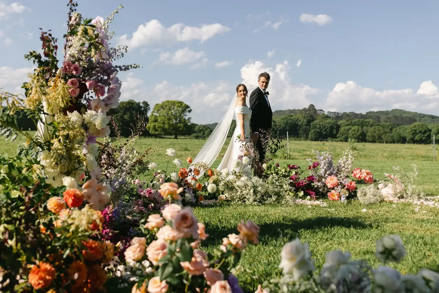 A bride and groom standing outdoors on their wedding day, surrounded by colorful flowers and a scenic green landscape under a blue sky with clouds.