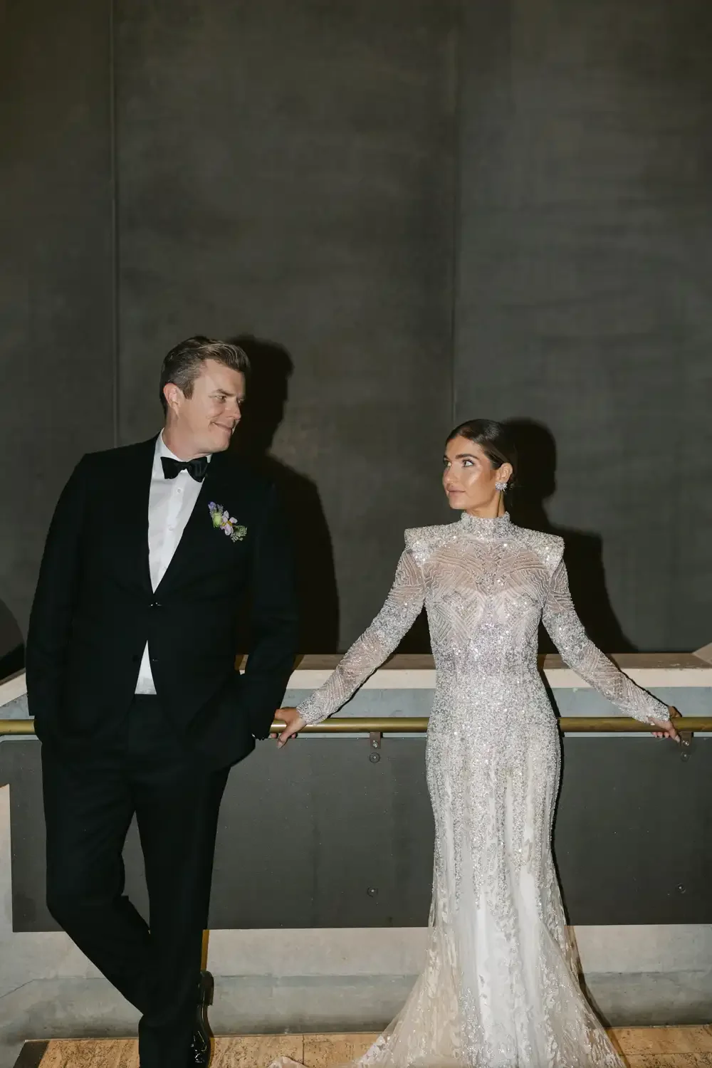 A man and woman, dressed in formal attire, are standing together. The man is wearing a black tuxedo with a white shirt and black bow tie, and the woman is in an elegant, long, sparkling gown. They are holding hands in front of a dark background, look