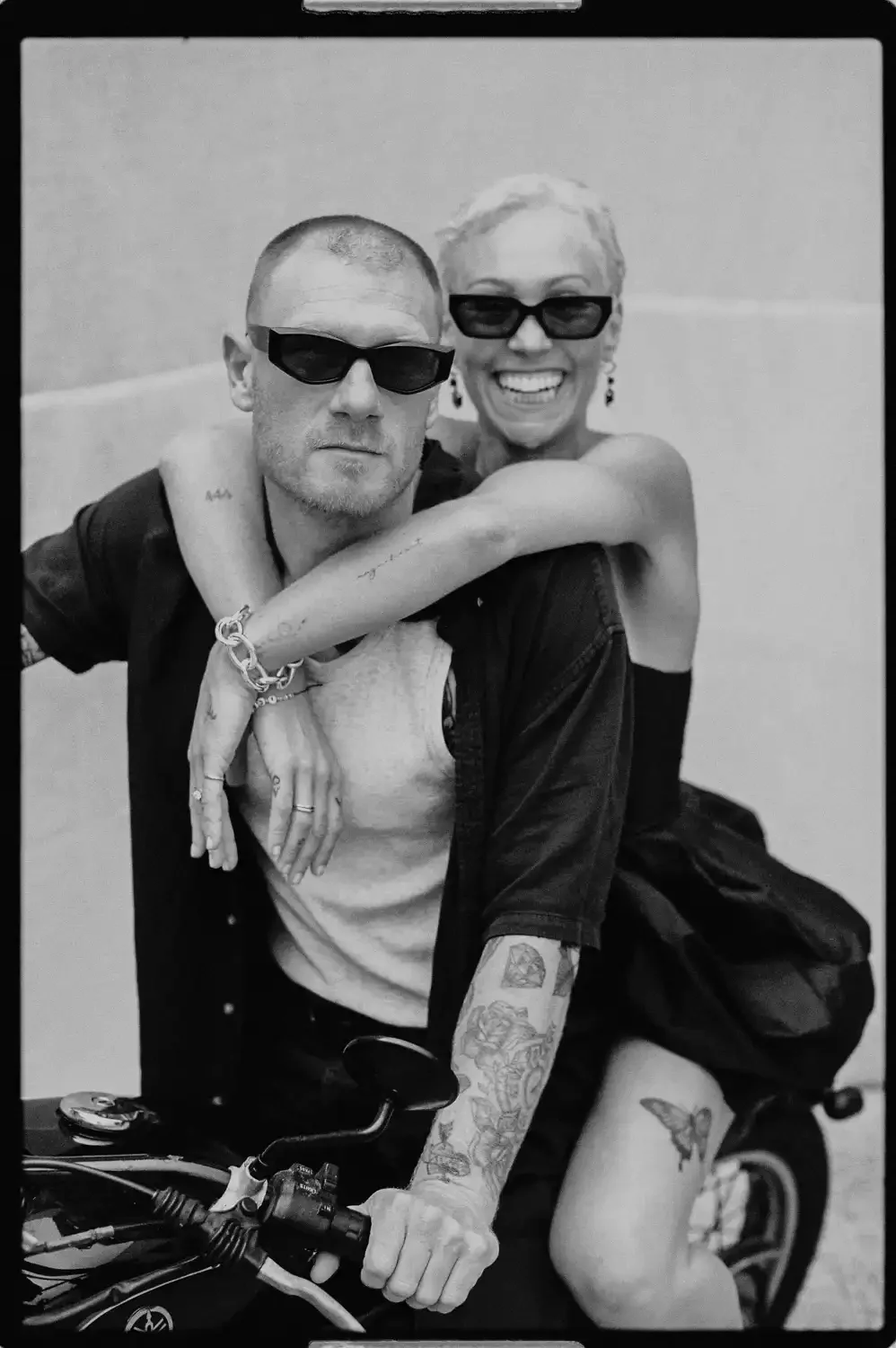 Black and white photo of a man with tattoos on his arm and a woman with short blonde hair and glasses, riding a motorcycle. The woman is hugging the man from behind and smiling.