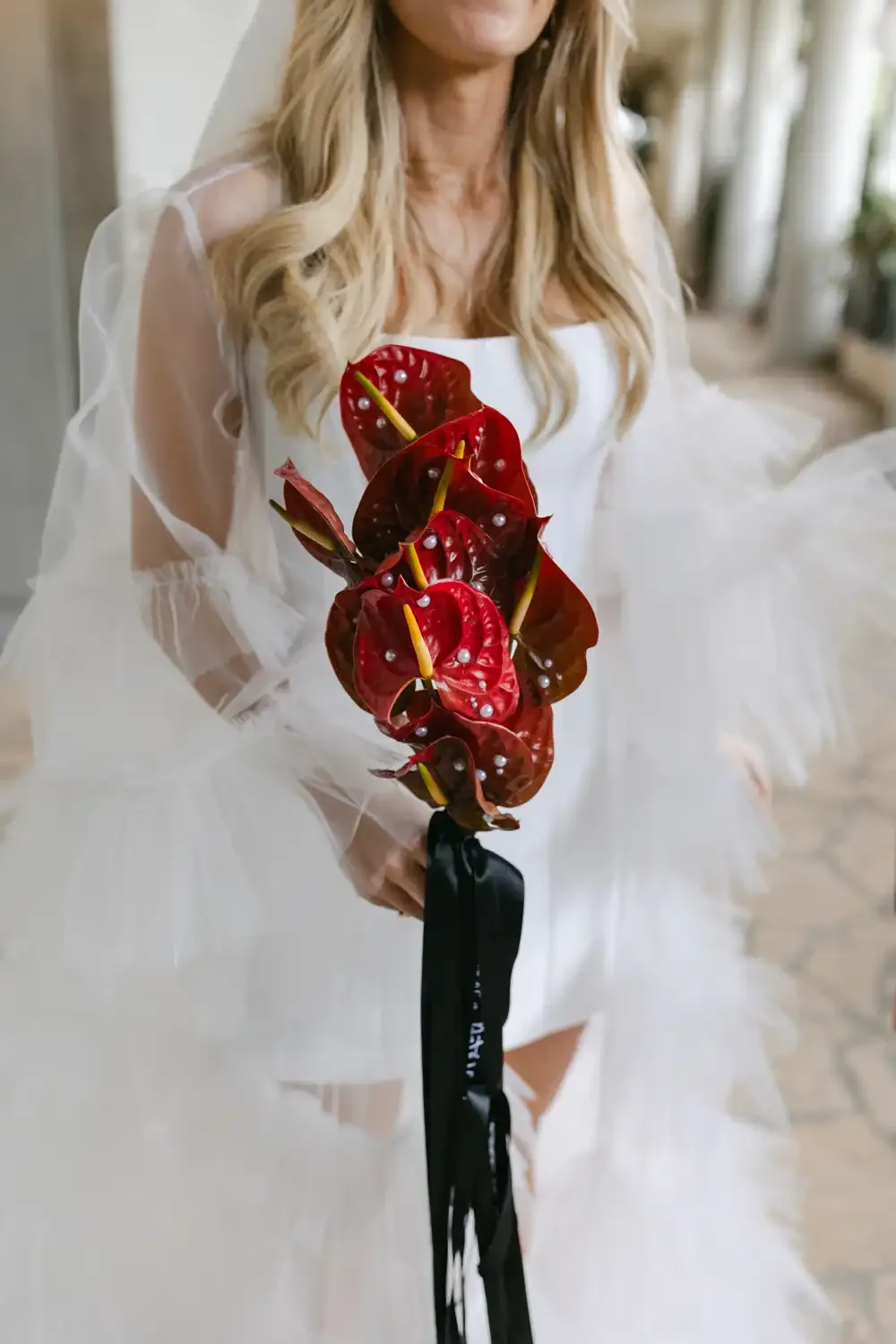 A woman in a white dress holding a bouquet of red anthurium flowers with pearls on the petals, tied with a black ribbon.