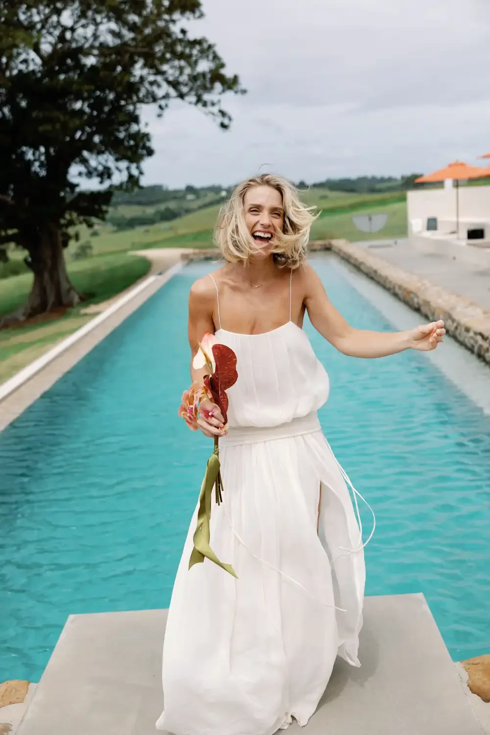 A woman in a white dress holding a flower, smiling by a swimming pool with a scenic background.
