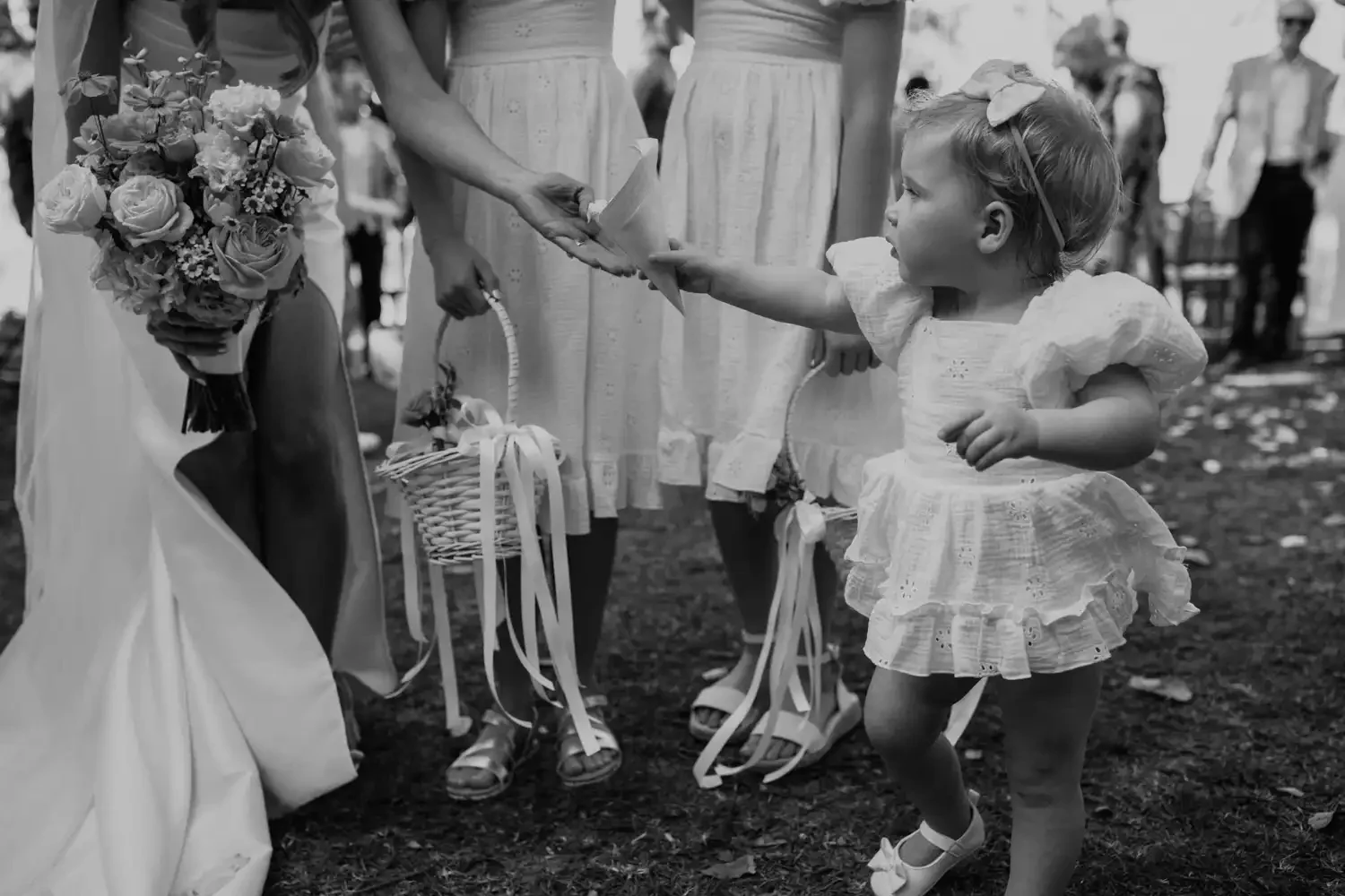 A young girl in a white dress reaching out to touch a woman holding a bouquet of flowers at an outdoor event.
