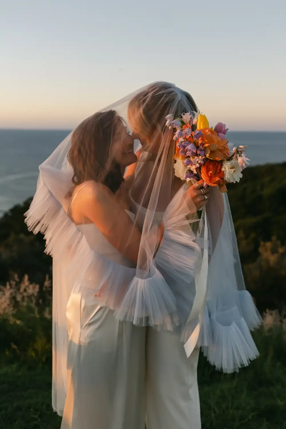 Two women in wedding dresses sharing a kiss outdoors during sunset, one woman holding a colorful bouquet, with ocean and greenery in the background.