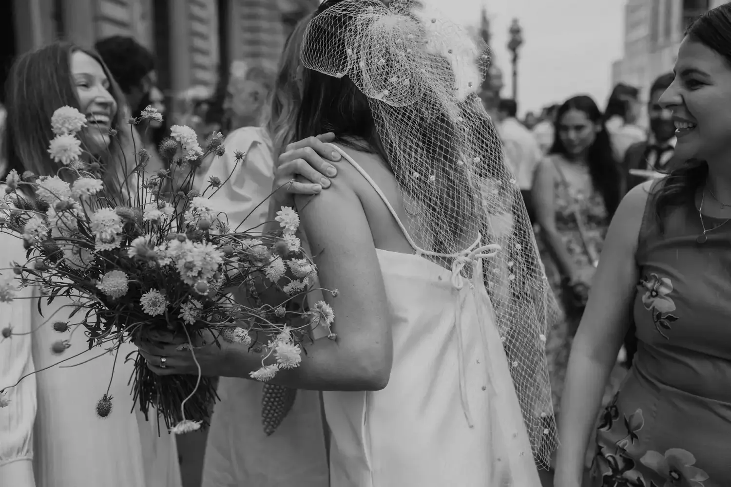A woman wearing a fascinator with netting receives a bouquet of flowers from another woman during a celebration or event, with smiling people in the background.