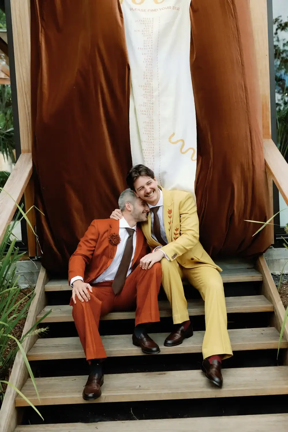 Two men dressed in colorful suits sitting on stairs, laughing and leaning toward each other, in front of a large, decorative backdrop with a list of names and a snake graphic, surrounded by plants.
