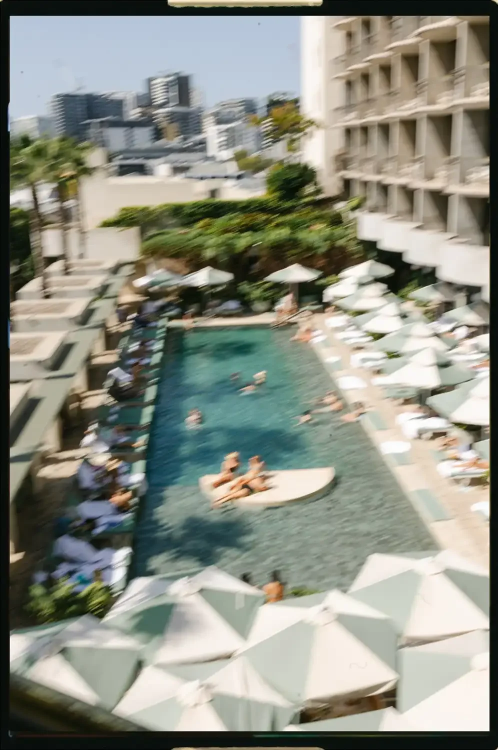 People swimming and relaxing in a hotel pool area with lounge chairs, umbrellas, and surrounding high-rise buildings.