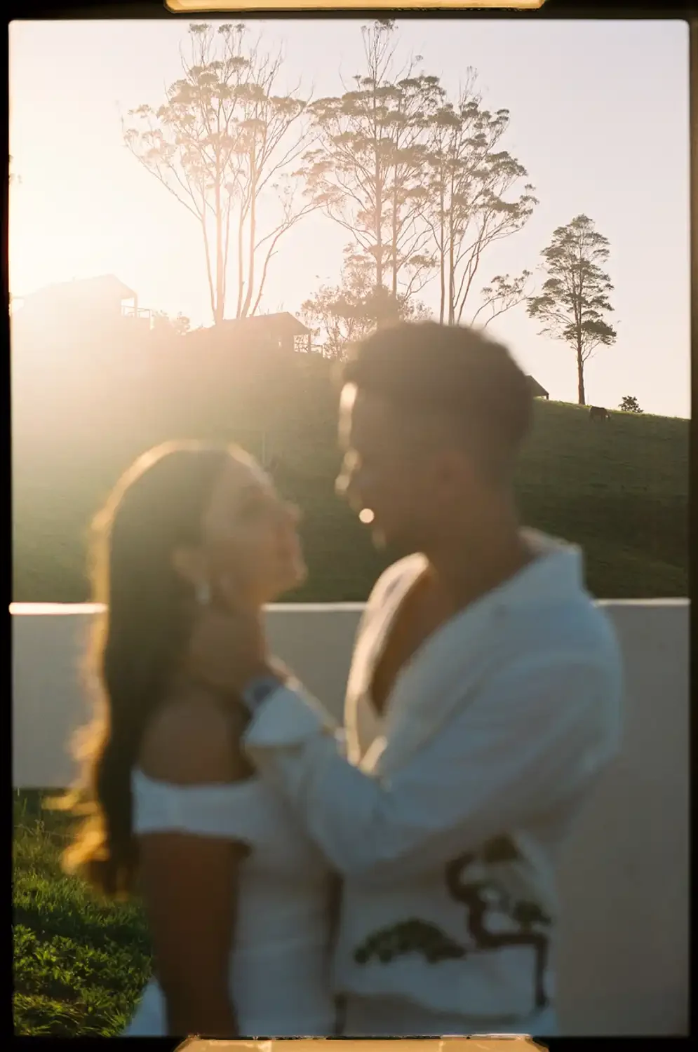A silhouetted couple facing each other during sunset, with trees and hills in the background.