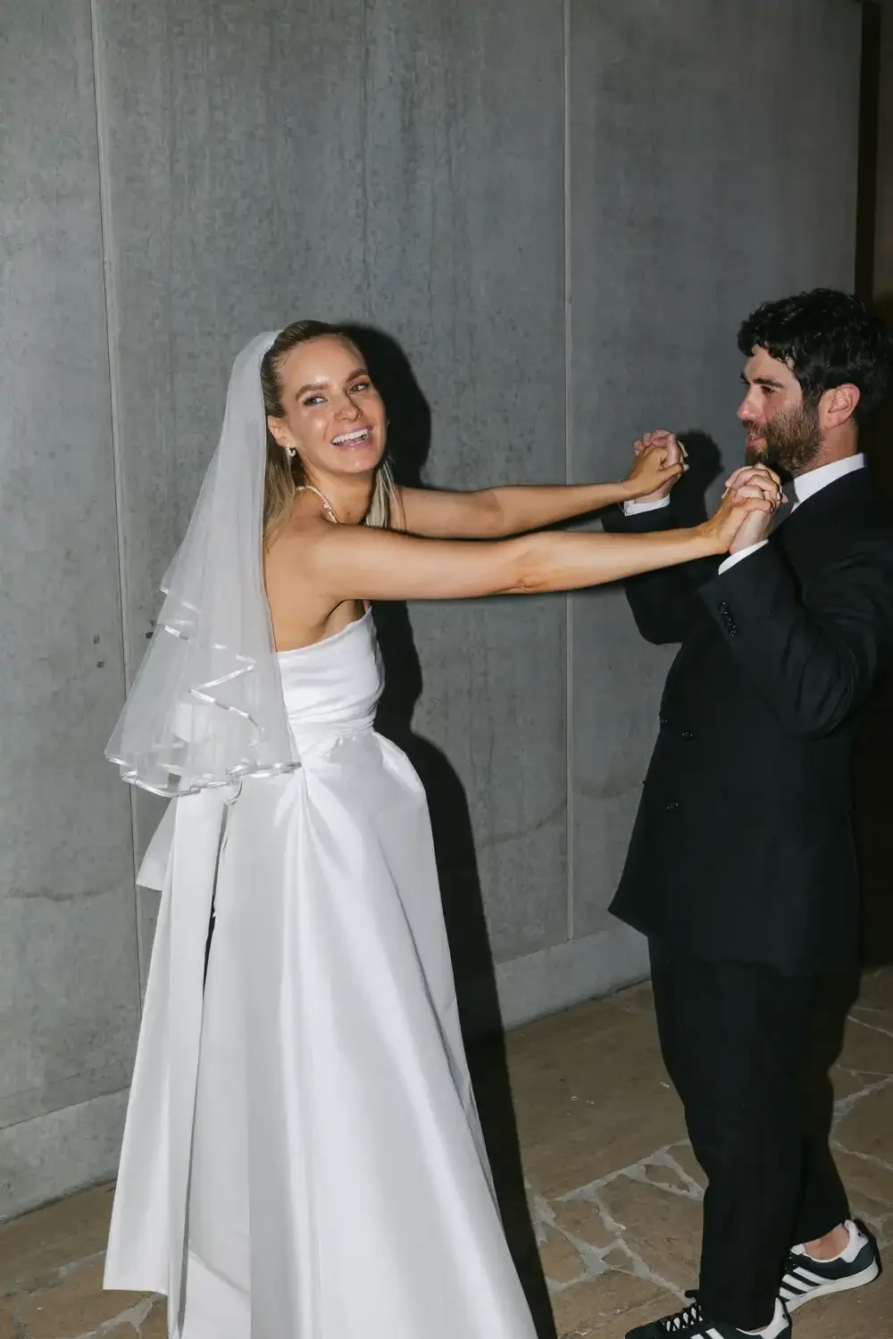 A bride and groom are dancing together in a playful manner at their wedding. The bride is pulling the groom's hands while smiling, and she is wearing a white wedding dress and veil. The groom is dressed in a black suit with a white shirt and black sn