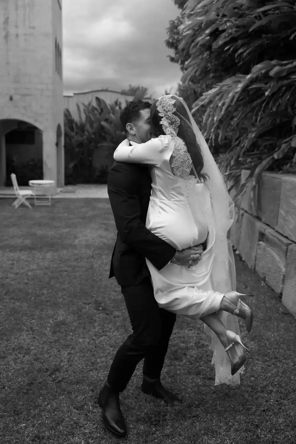 A man in a black suit lifting a woman in a white wedding dress with a lace veil, both are hugging and appear to be celebrating outdoors with large plants and a stone building in the background.
