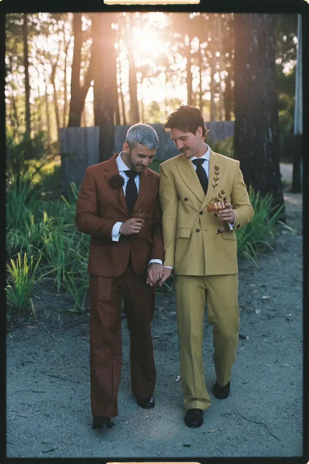 Two men in suits walking arm in arm outdoors during sunset, holding hands, with greenery and trees in background.
