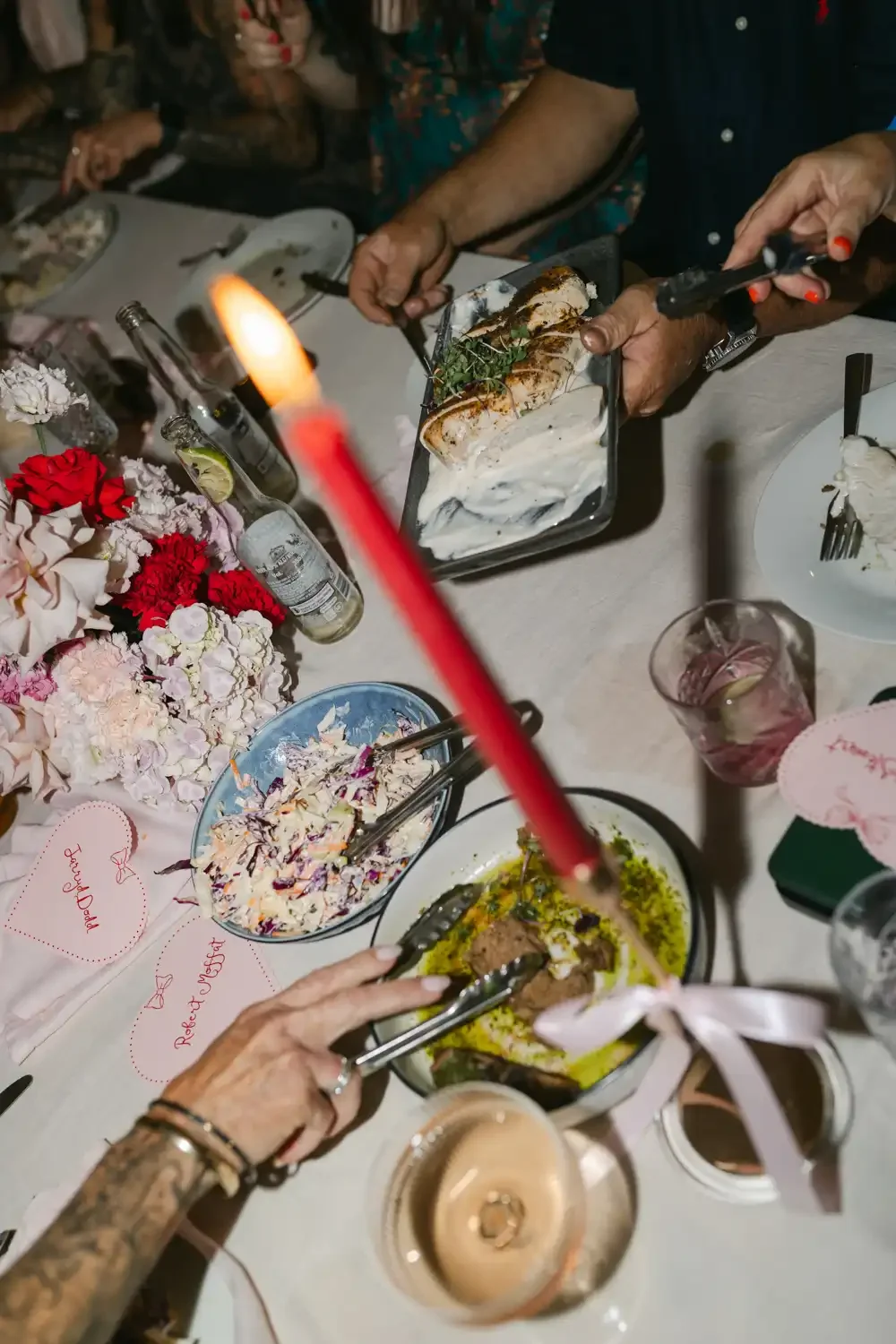 People gathered around a dining table with flowers and drinks, serving themselves various dishes, including a large baked fish with herbs, a bowl of salad, and other foods.
