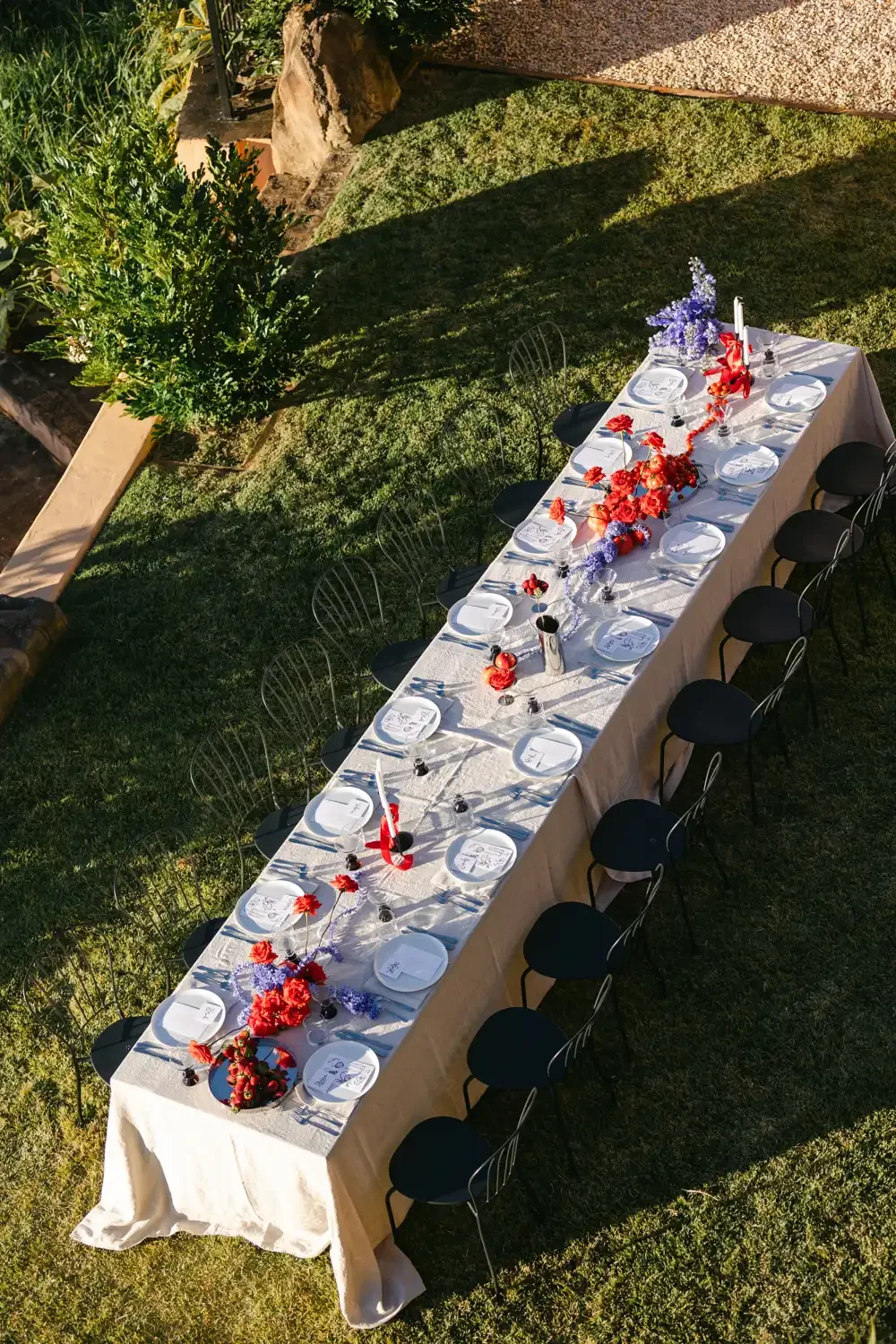 Long outdoor dining table decorated with red and purple flowers, tied with red ribbons, set with white plates, glasses, and cutlery. Surrounded by black chairs on a grassy lawn, with a backdrop of trees and rocks.