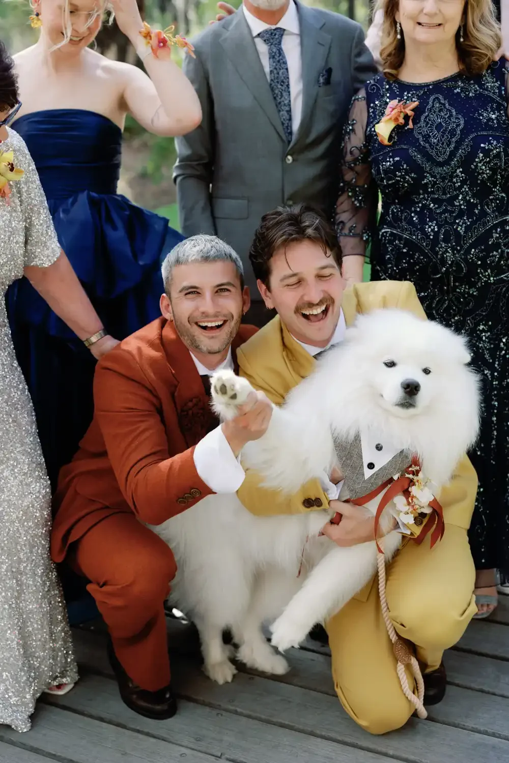 Group of people, including two men in bright-colored suits, holding a large white fluffy dog dressed in a tuxedo-style costume, smiling and celebrating outdoors.