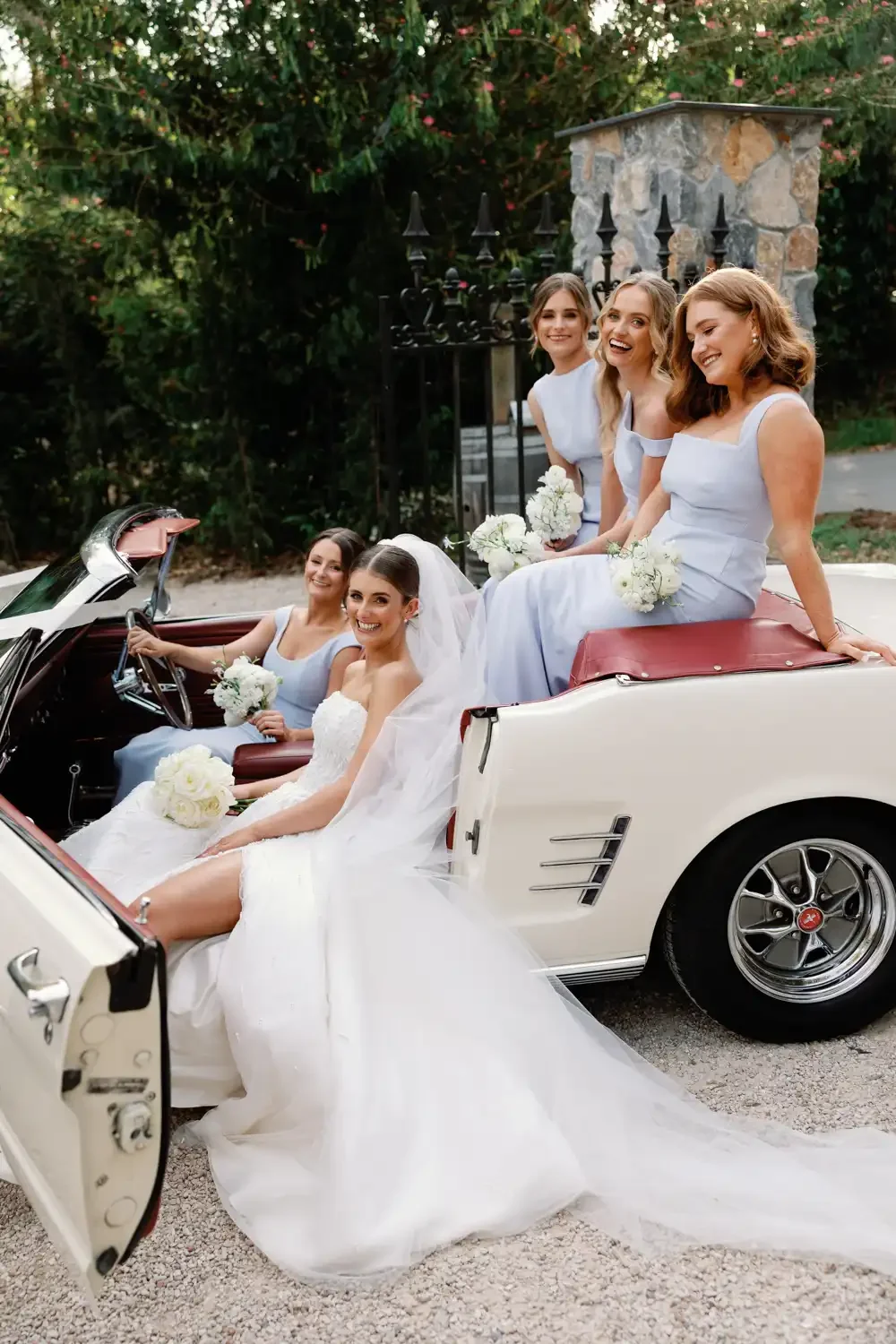 A bride in a wedding dress and veil sitting in a vintage white convertible car with five bridesmaids wearing matching light blue dresses, all holding white bouquets, outdoors with greenery and a stone structure in the background.
