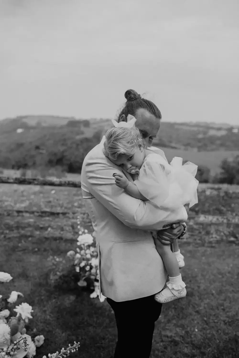 A man with a bun hairstyle holding a young girl in an outdoor setting, flowers in the background, black and white photograph.