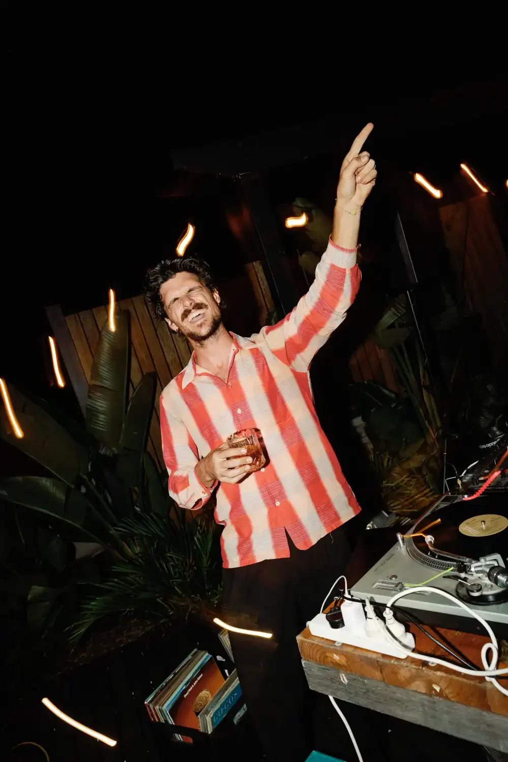 A man in a red and beige checkered shirt holding a drink and dancing at a party or DJ event.