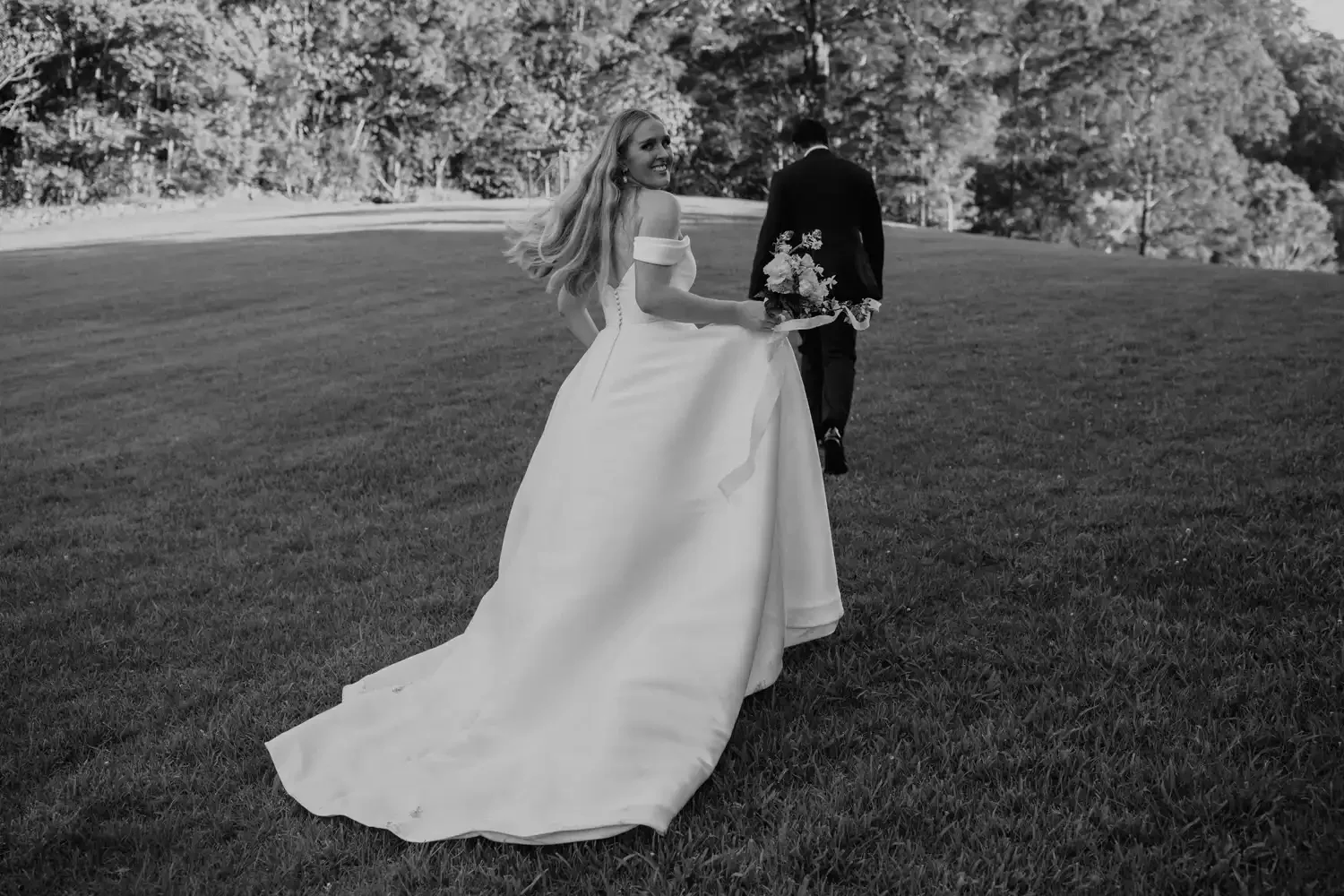 A bride holding a bouquet, smiling, walking on a grassy field with a man in a suit walking ahead, surrounded by trees.