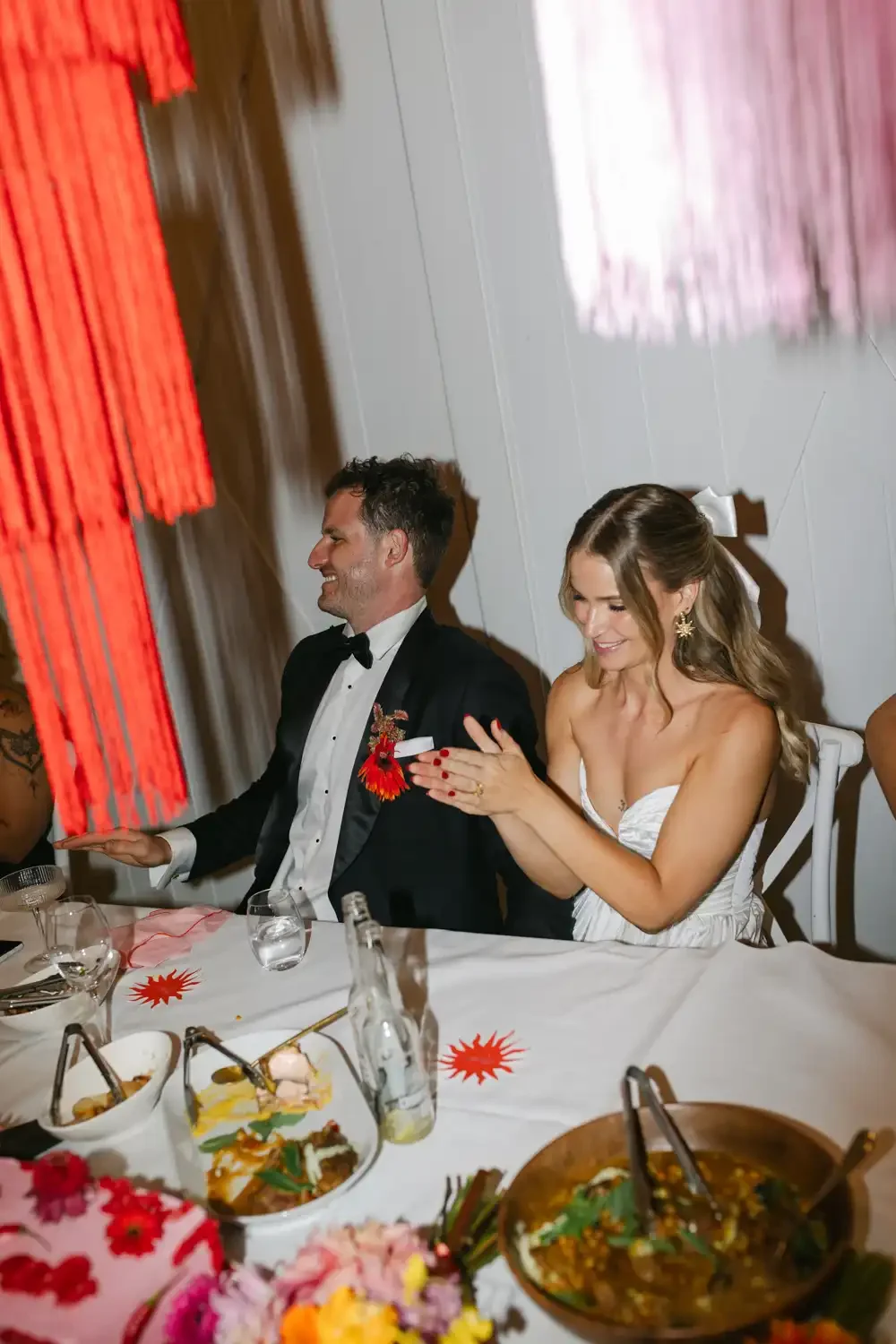 A man in a tuxedo and a woman in a white dress are sitting at a wedding reception table, smiling and clapping. They are decorated with flowers and surrounded by food and drinks.