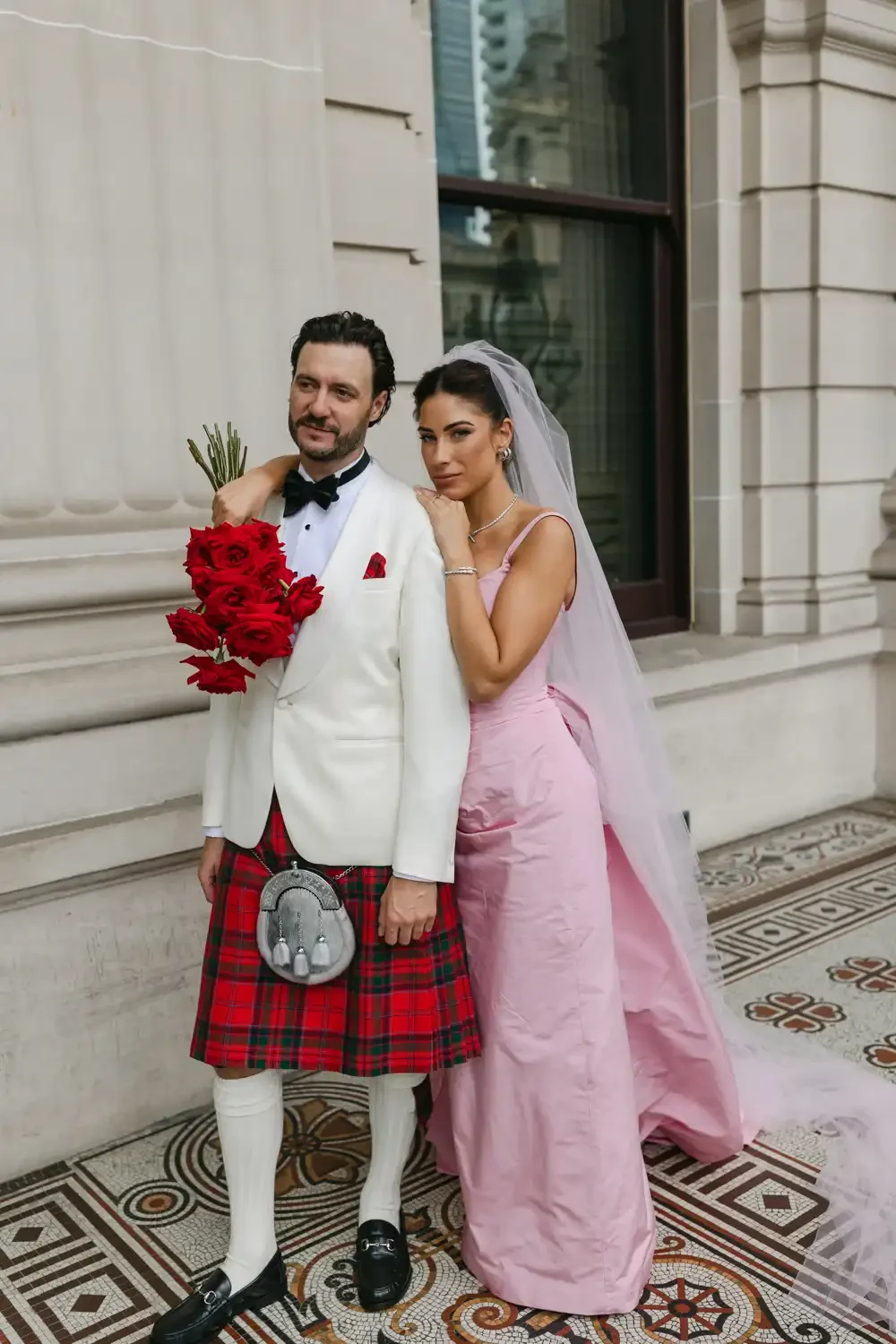 A bride and groom dressed in wedding attire standing outside a building with stone walls and large windows. The groom is wearing a white tuxedo jacket, red tartan kilt, and holding a bouquet of red roses. The bride is in a pink gown with a veil, rest