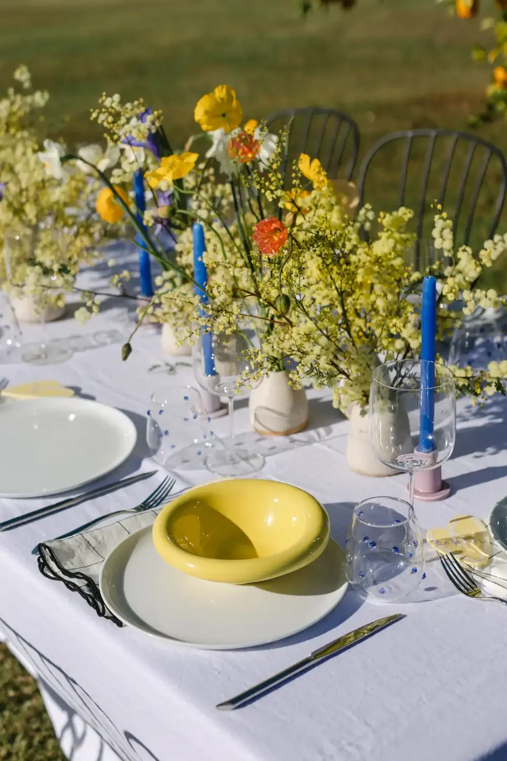 A beautifully arranged outdoor table setting with white tablecloth, yellow and white flower centerpieces, blue candles, and colorful dishes and glasses.