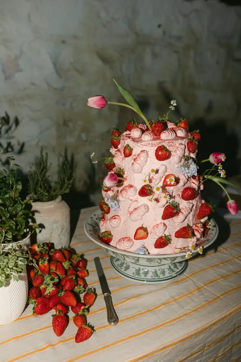 A pink, layered strawberry cake decorated with fresh strawberries, small white flowers, tulips, and greenery, placed on a decorative cake stand on a table with a striped tablecloth, with potted plants nearby.