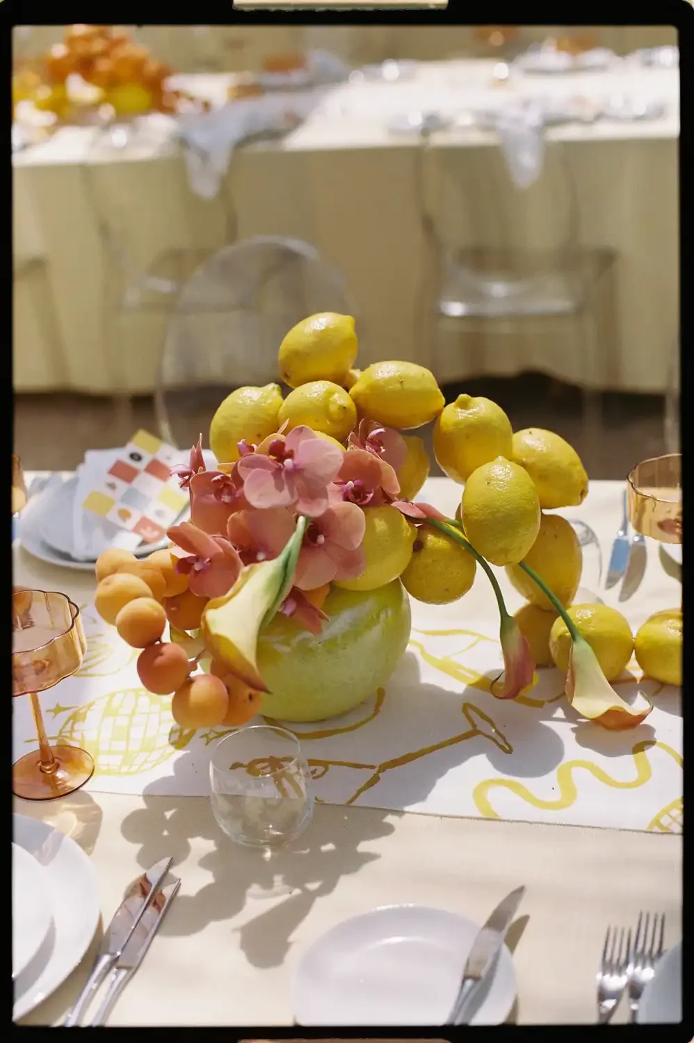 Table centerpiece made of lemons, peach-colored flowers, and peach-colored berries in a green bowl, set on a white tablecloth with gold patterns, surrounded by glassware and cutlery.