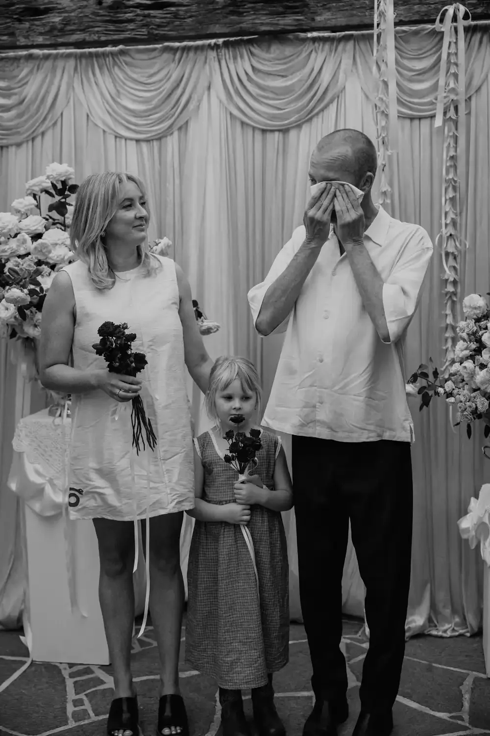 A family at a celebration, with a woman holding flowers, a girl also holding flowers, and an emotional man wiping his eyes, standing in front of decorated curtains and floral arrangements.