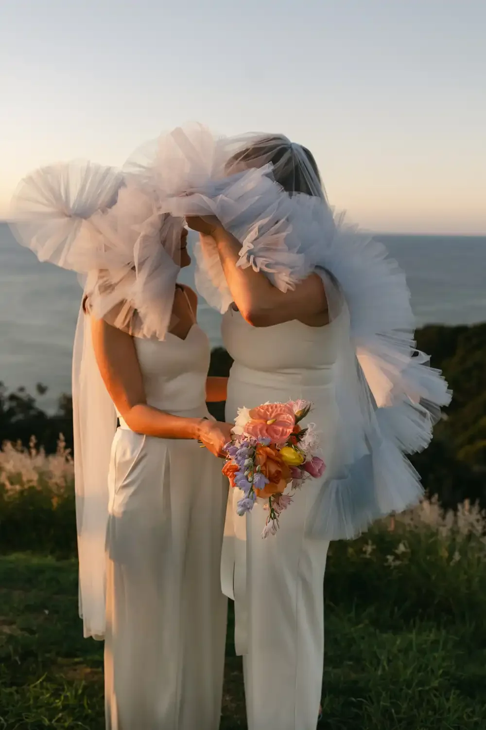 Two women in white dresses, wearing large white ruffled headpieces, stand close together outdoors at sunset, holding a bouquet of colorful flowers.