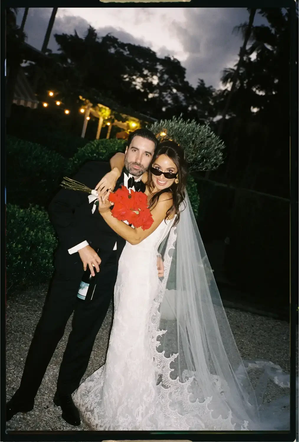 A bride and groom at a wedding, embracing outdoors at dusk. The bride wears a white lace wedding gown with a long veil, and holds a bouquet of red flowers. The groom is dressed in a black tuxedo and is holding a bottle of wine.
