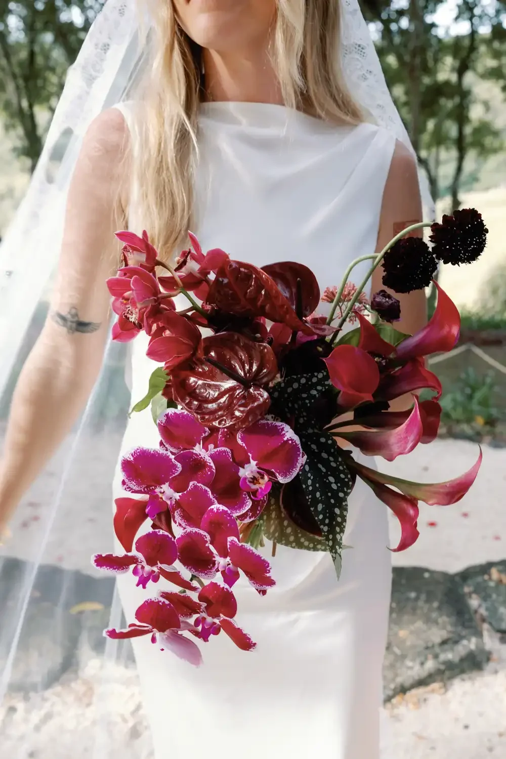 A woman in a white dress holding a bouquet of dark and pink flowers, standing outdoors with greenery in the background.