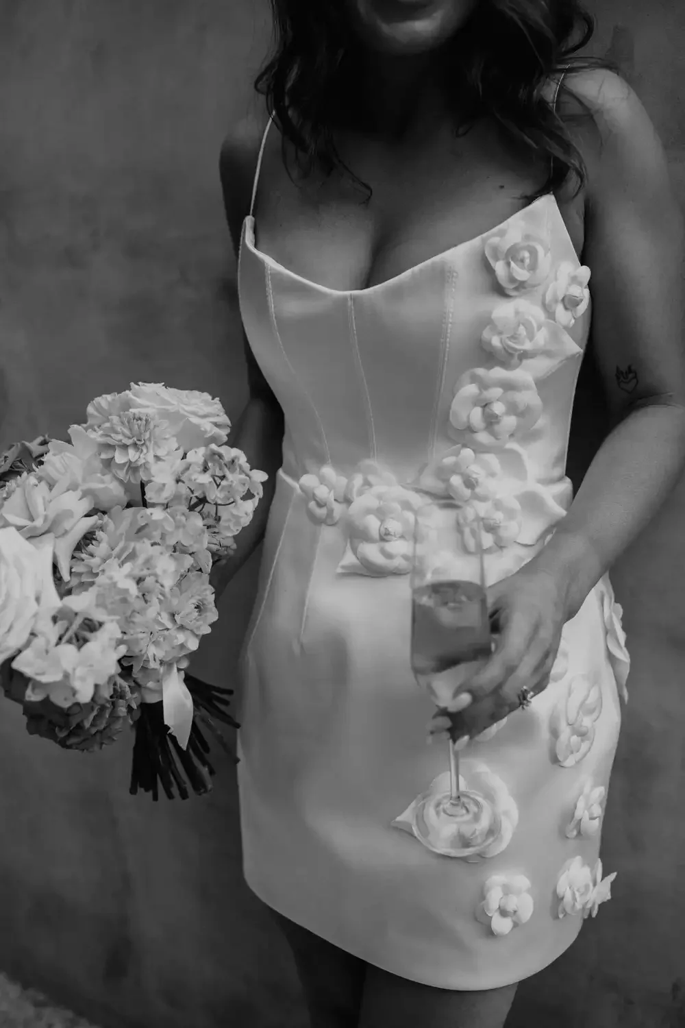 Woman in a white dress with floral appliqué, holding a bouquet of flowers in one hand and a glass of champagne in the other.
