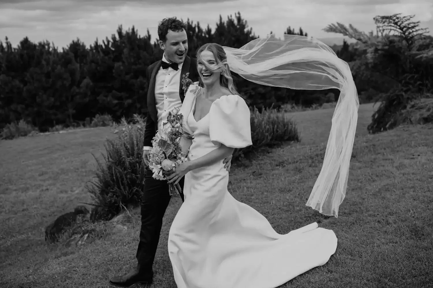 A smiling bride and groom on their wedding day outdoors, with the bride holding a bouquet of flowers and wearing a long, flowing wedding dress with puffed sleeves, and the groom in a tuxedo with a bowtie. The bride's veil is blowing in the wind, and 
