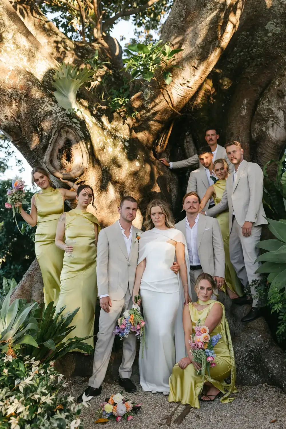 A group of people, including a bride in a white dress and several men in suits and women in yellow dresses, gathered around a large tree in a garden setting, possibly for a wedding photoshoot.