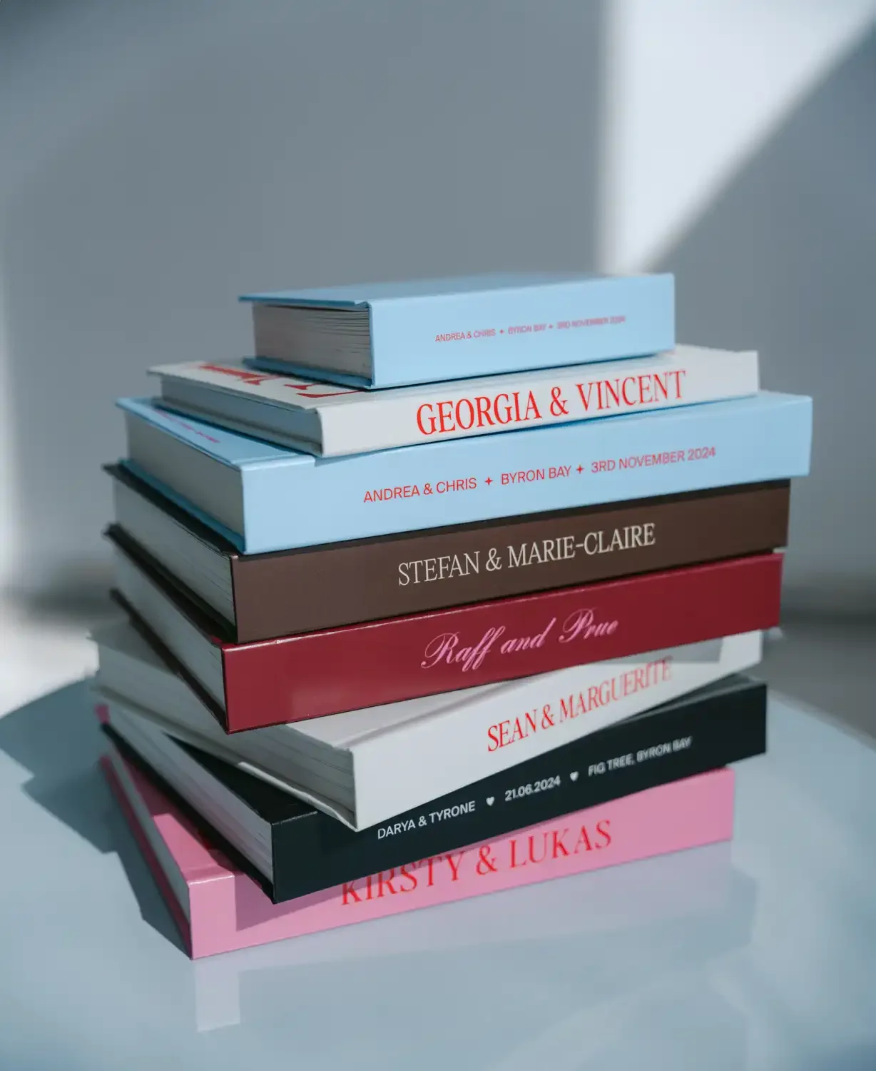 Stack of wedding invitation books with titles in various colors, including 'Georgia & Vincent,' 'Stefan & Marie-Claire,' and names like 'Raff and Pru,' arranged on a light surface with a shadow cast on the background.