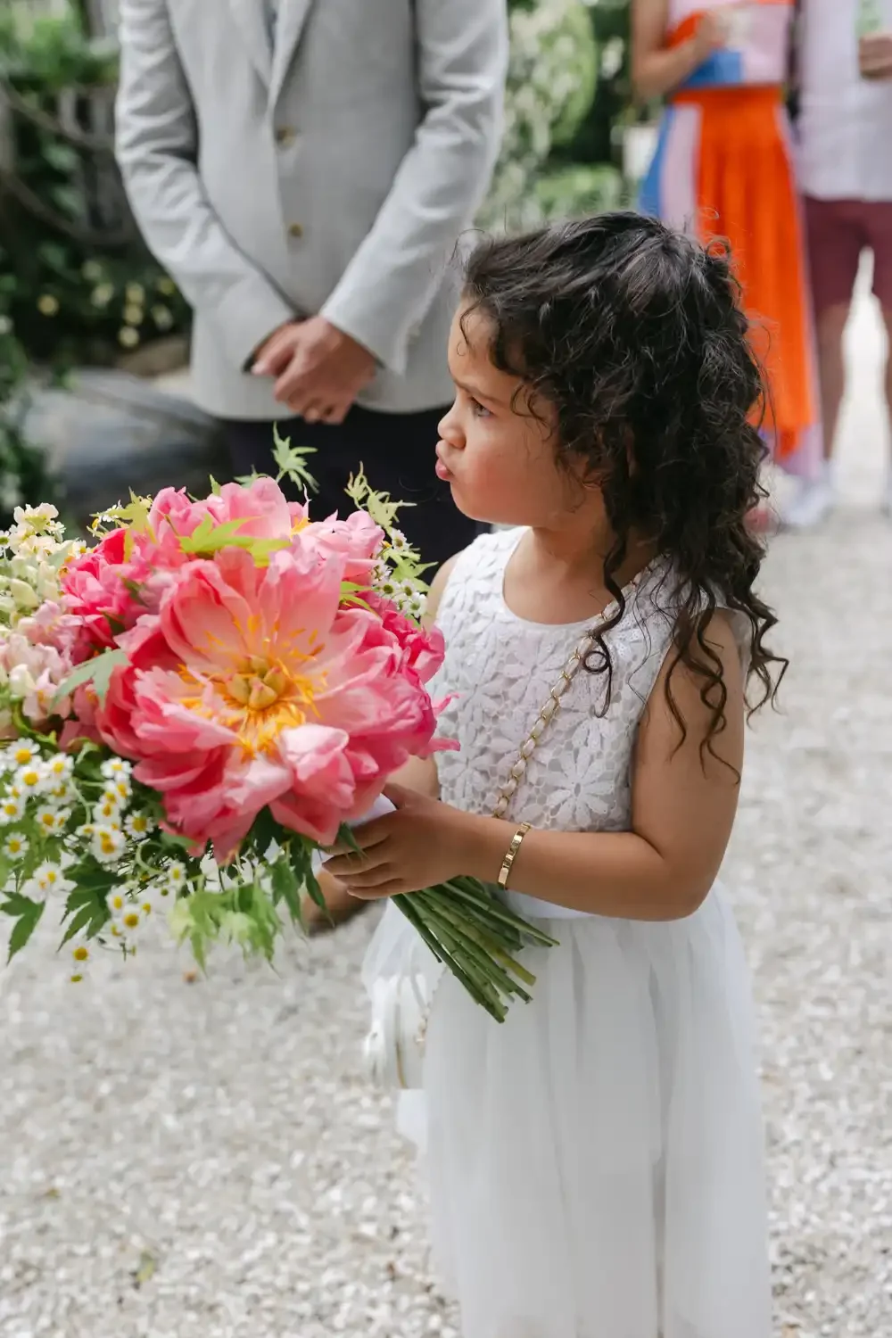 A young girl with curly hair dressed in a white dress holding a large bouquet of pink and yellow flowers, standing outdoors during a formal event with adults in background.