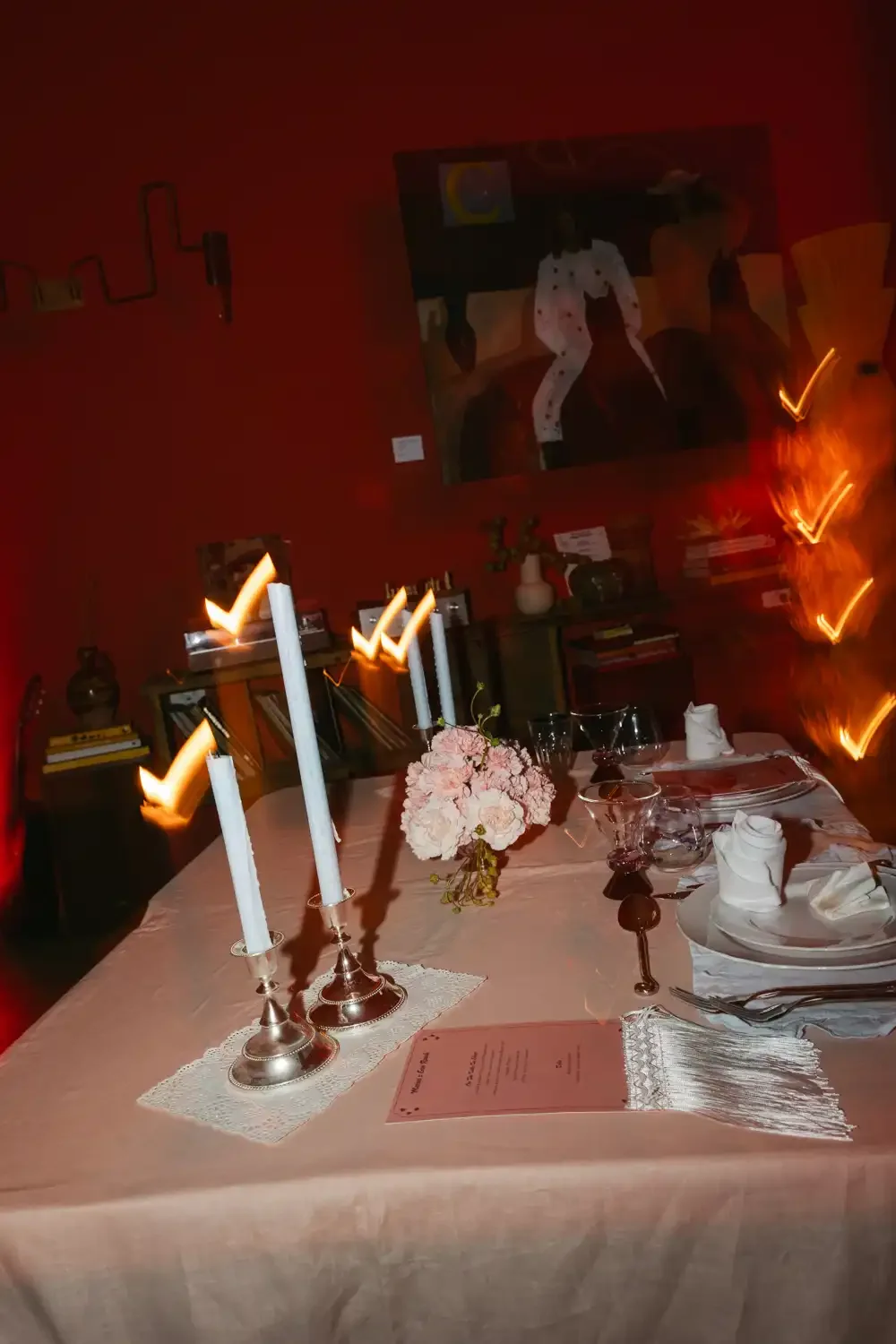 A dining table set for a formal dinner with white tablecloth, pink floral centerpiece, white napkins, wine glasses, and candlesticks with lit candles. The background features a red wall with a painting and bookshelves.