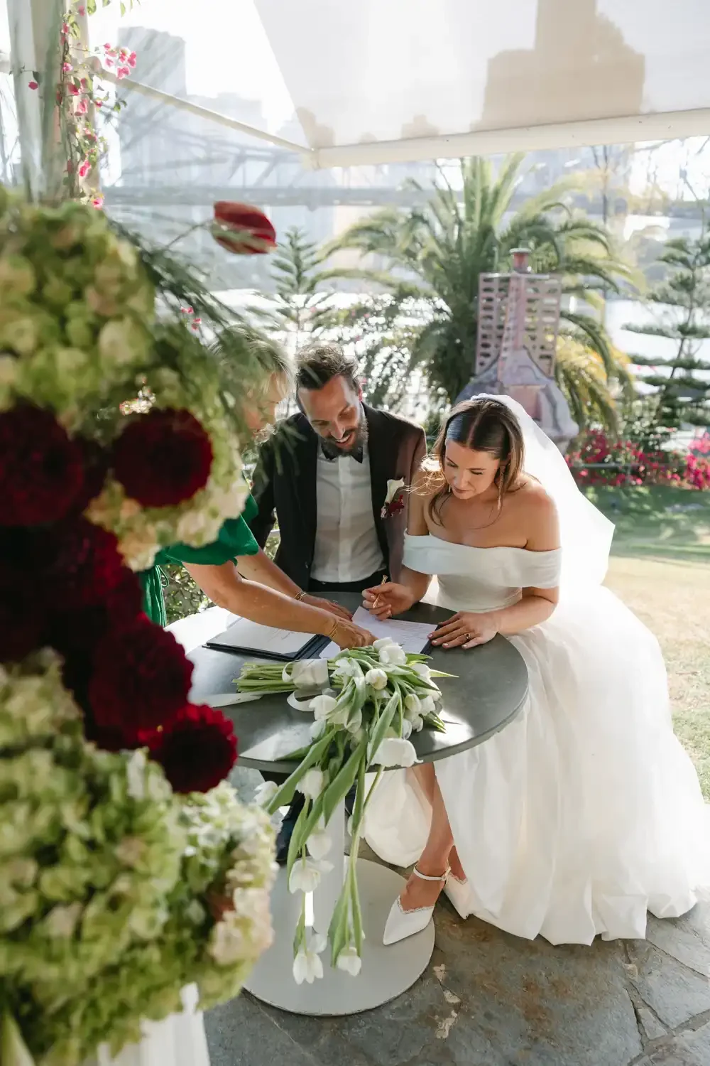 A bride and groom signing marriage documents at an outdoor wedding ceremony with a floral table and lush greenery in the background.