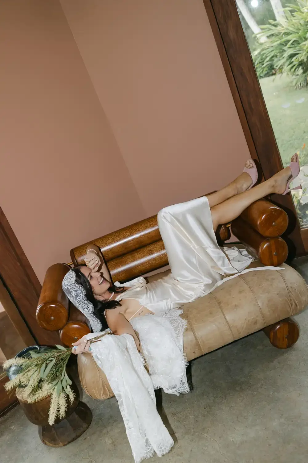 A woman in a wedding dress lying on a wooden salon-style sofa, smiling, holding a bouquet, wearing a lace veil and high heels, in a room with a large window showing greenery outside.