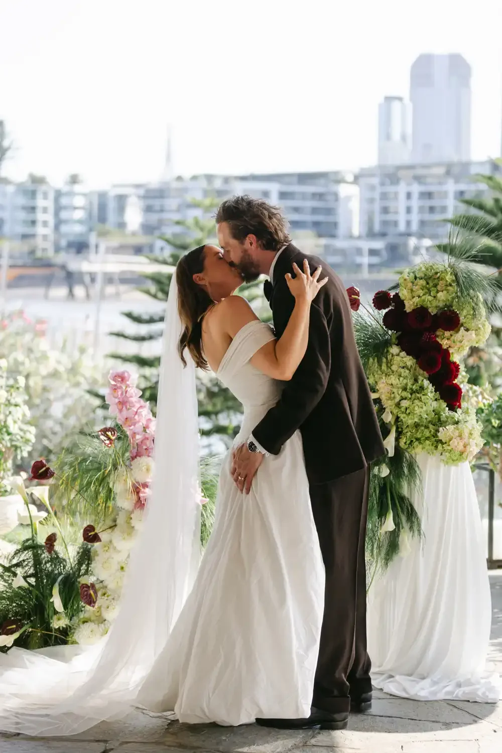 Bride and groom sharing a kiss at their wedding ceremony with cityscape backdrop and floral arrangements.