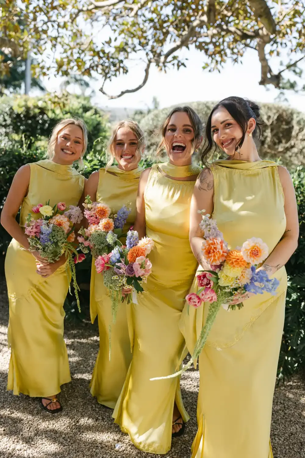 Four women in yellow dresses holding colorful bouquets, standing outdoors under trees, smiling and posing for a photo.