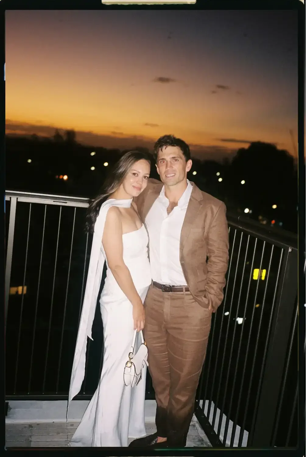 A woman and a man stand close together on a balcony during sunset. The woman is wearing a white dress and holding a white purse, while the man is wearing a brown suit and white shirt. They are smiling and looking at the camera against a backdrop of a