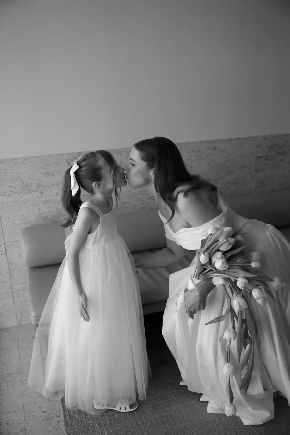 A woman dressed in a gown kissing a young girl in a white dress, both leaning in for a kiss, in a tender moment, with the woman holding a bouquet of tulips.