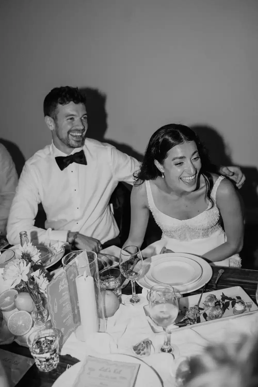 A black and white photo of a man and woman smiling and laughing at a dinner table during a celebration. The man is wearing a white shirt and black bow tie, and the woman is in a sleeveless dress with beaded details. The table is decorated with flower