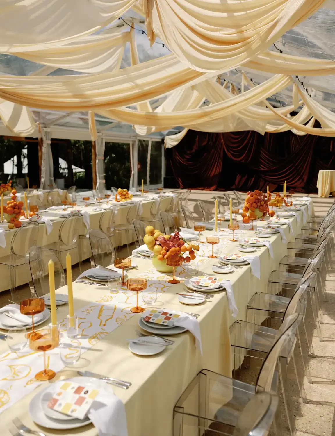 An indoor banquet setup with a long table decorated with orange flowers and candles, transparent chairs, draped cream-colored ceiling fabric, and a dark curtain backdrop.