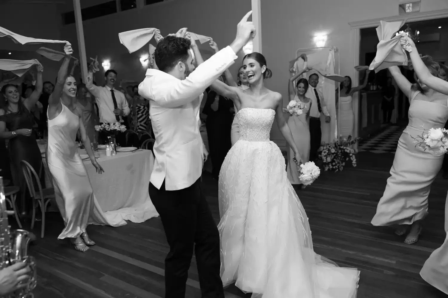 Black and white photo of a wedding reception with the bride and groom dancing in the center, surrounded by guests holding cloths in the air.